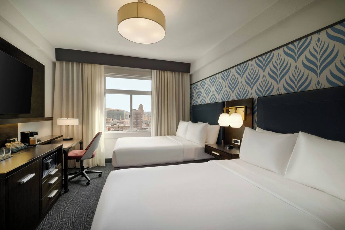 Habitación de hotel con dos camas, un escritorio, una silla, un televisor y una ventana con vista a la ciudad.