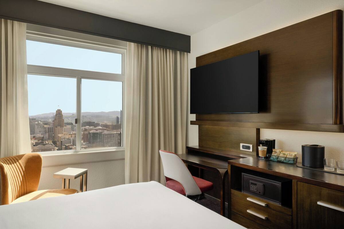Habitación de hotel con una cama grande, un escritorio, un televisor de pantalla plana, una silla, cafetera y vistas a un paisaje urbano a través de un ventanal amplio.