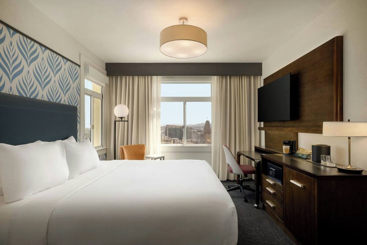 Habitación de hotel con una cama grande, un escritorio con silla, televisor y vista a la ciudad desde la ventana.