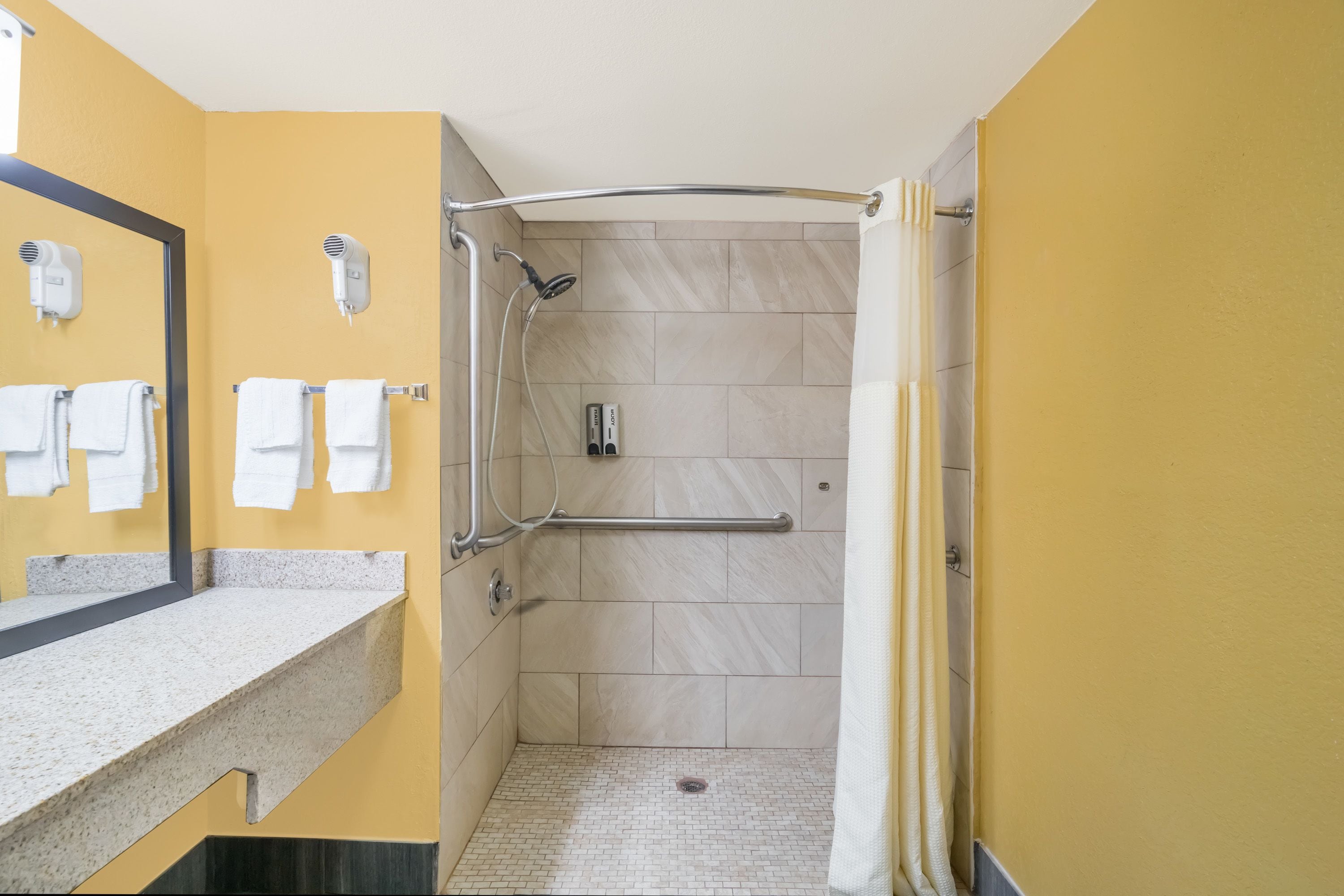 Un baño con pared amarilla, un espejo, un lavamanos, una ducha con barra de apoyo y dos secadores de pelo montados en la pared.