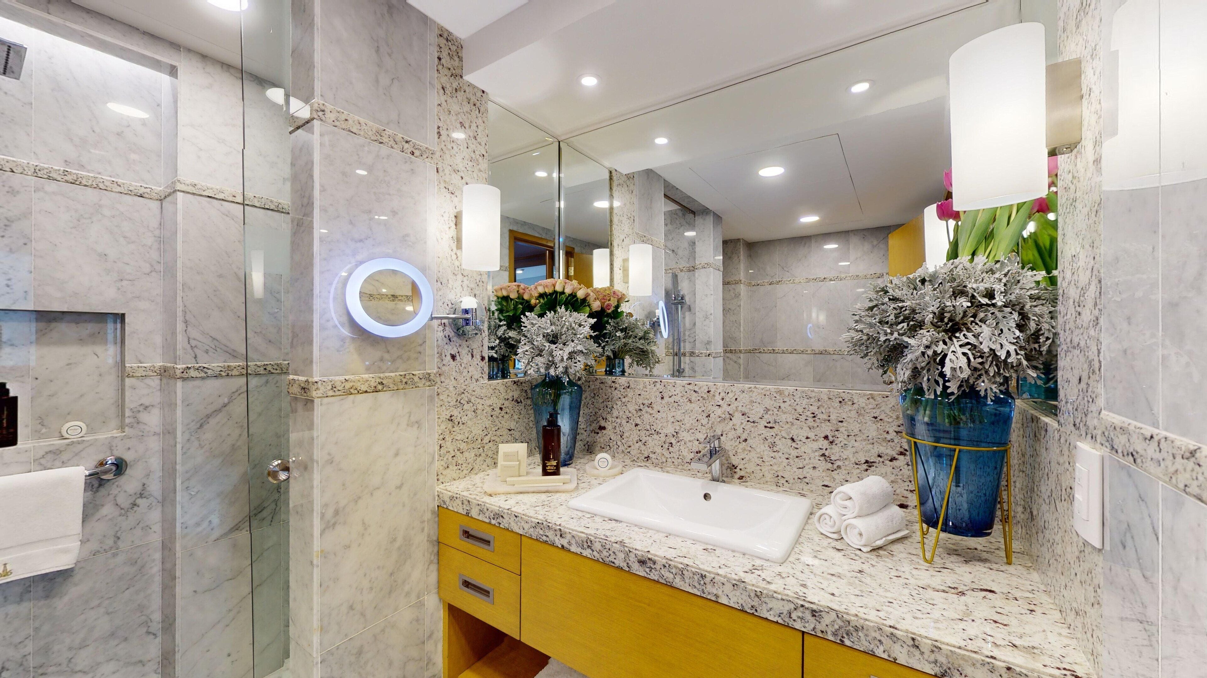 A modern bathroom with a marble vanity, a large mirror, and two flower arrangements.