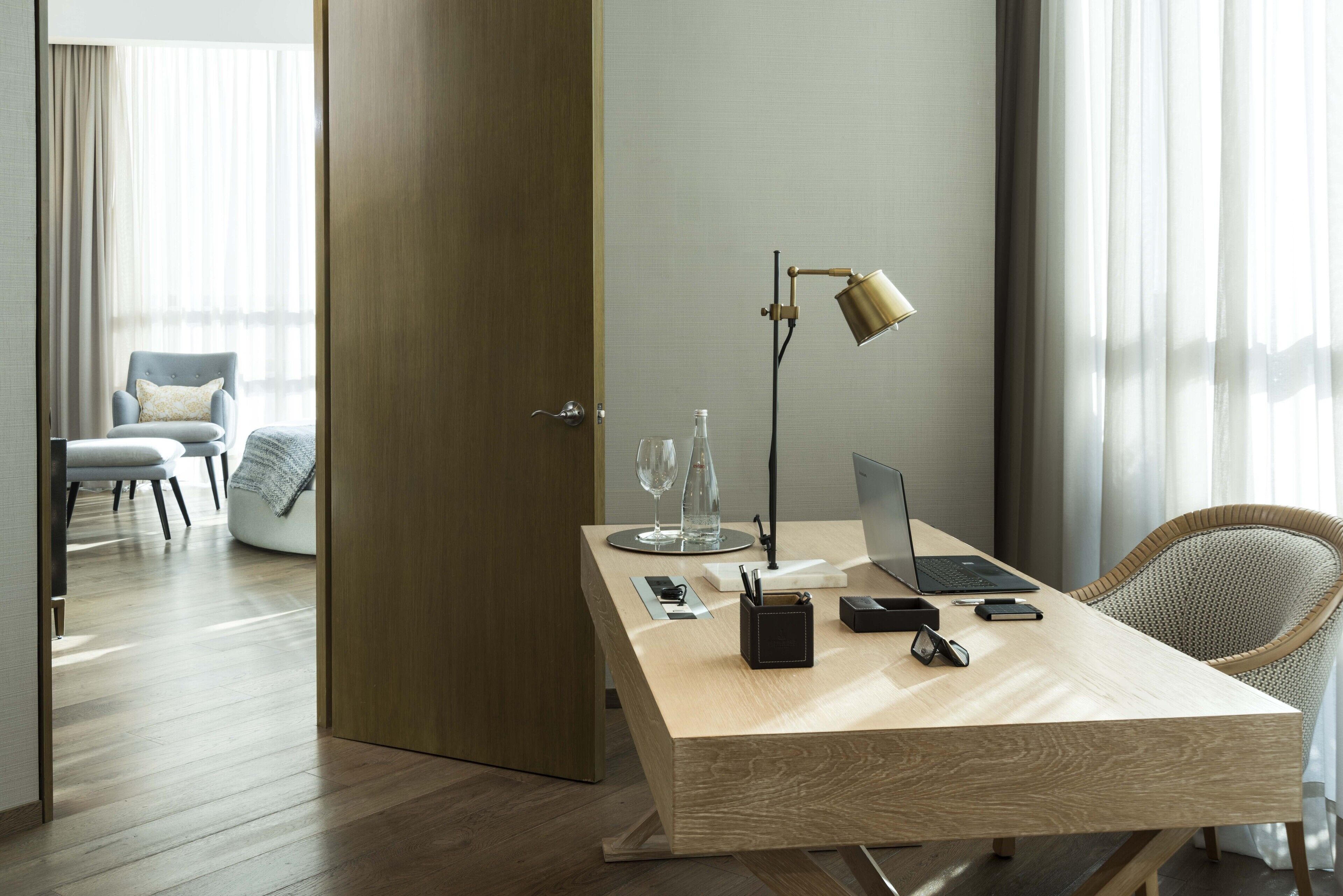 A modern hotel room with a wooden desk, laptop, and a lamp. There is a chair and a bed in the background.