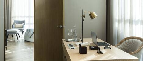 A modern hotel room with a wooden desk, laptop, and a lamp. There is a chair and a bed in the background.