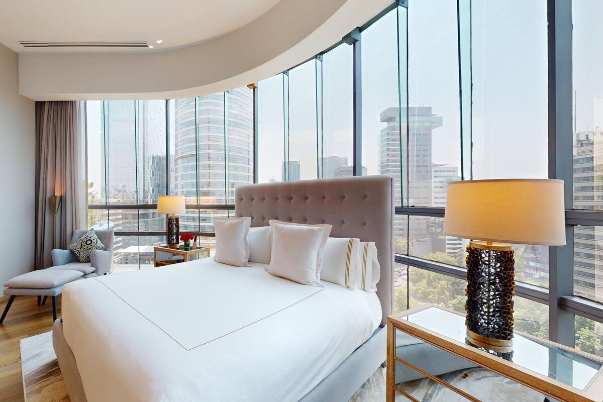 A modern hotel room with a large bed, a sitting area, and a view of the city skyline through floor-to-ceiling windows.
