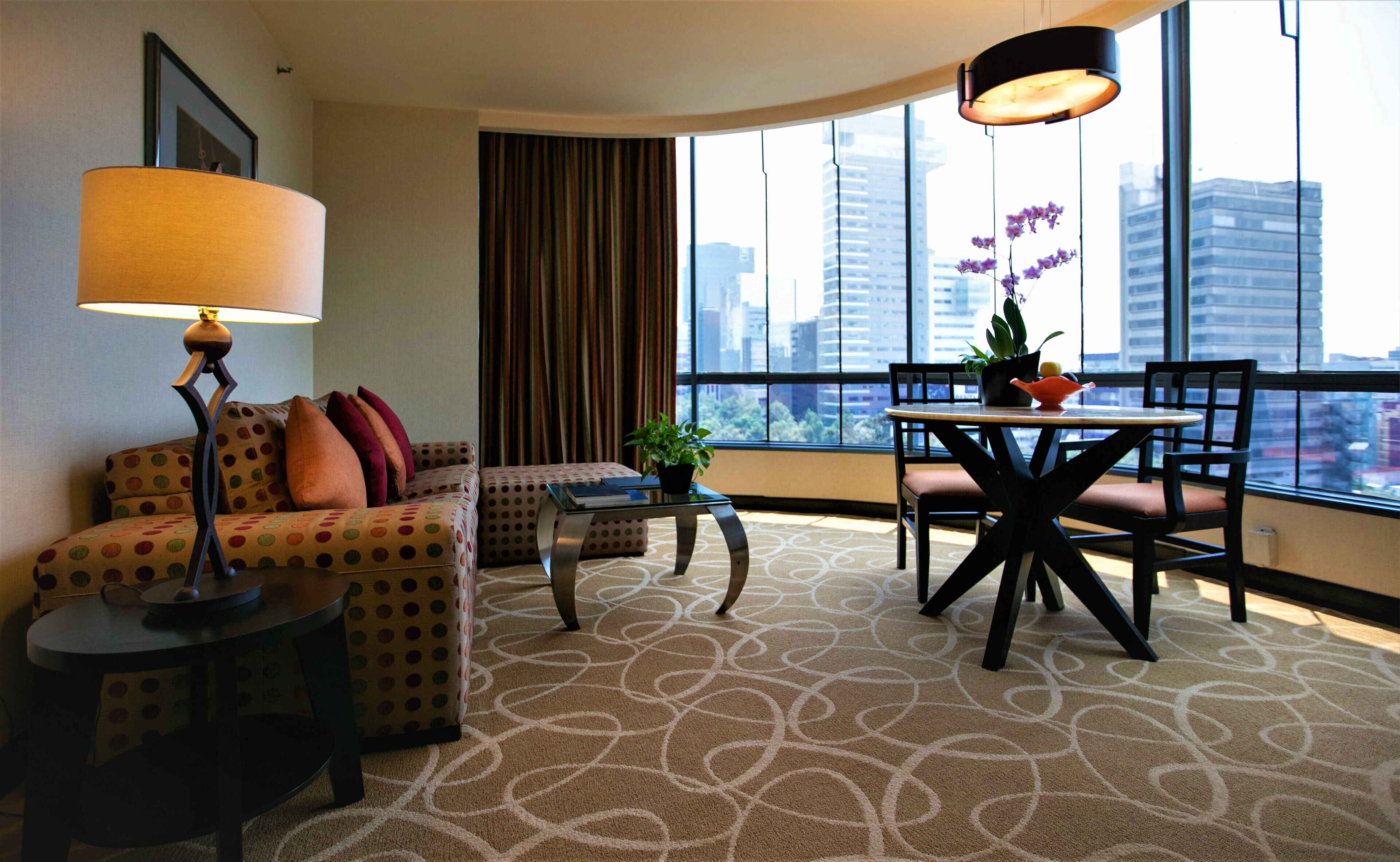 A hotel room with a sofa, a small table, a dining table with chairs, a lamp, a potted plant, and a view of the city through large windows.
