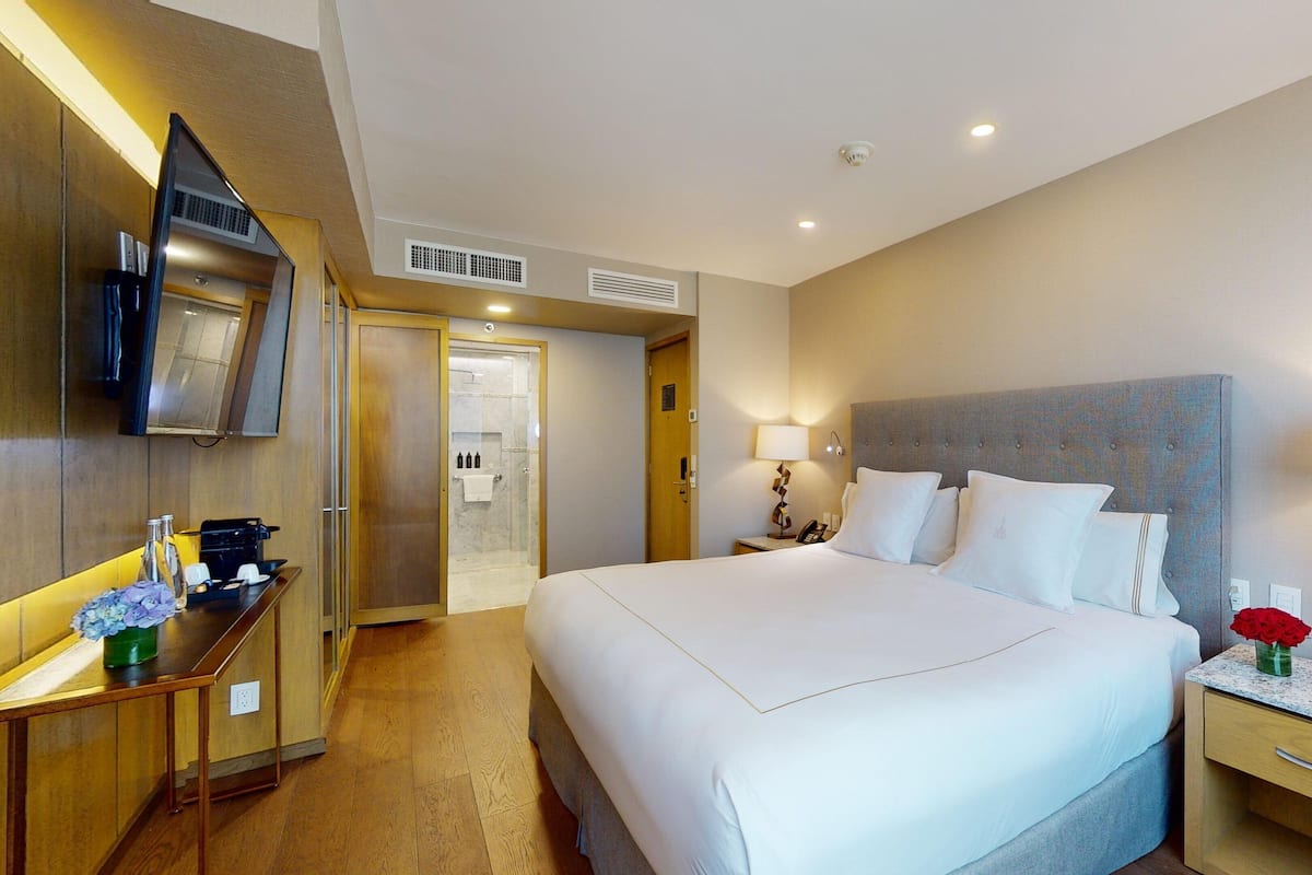 A hotel room with a large bed, bedside tables, a TV, a shower, and a desk with a coffee machine.