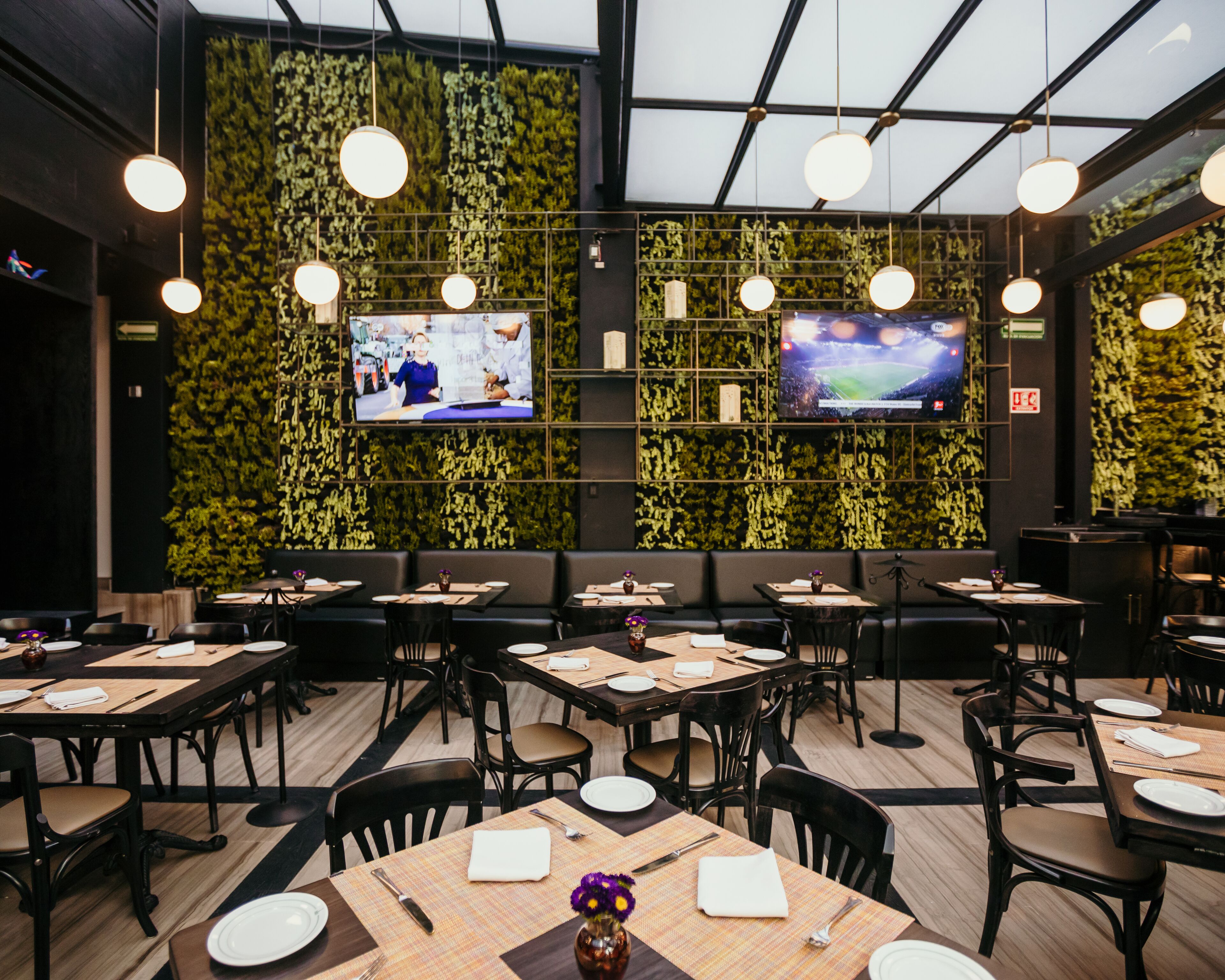 A restaurant with multiple tables and chairs, a green wall with plants, and two TVs mounted on the wall.
