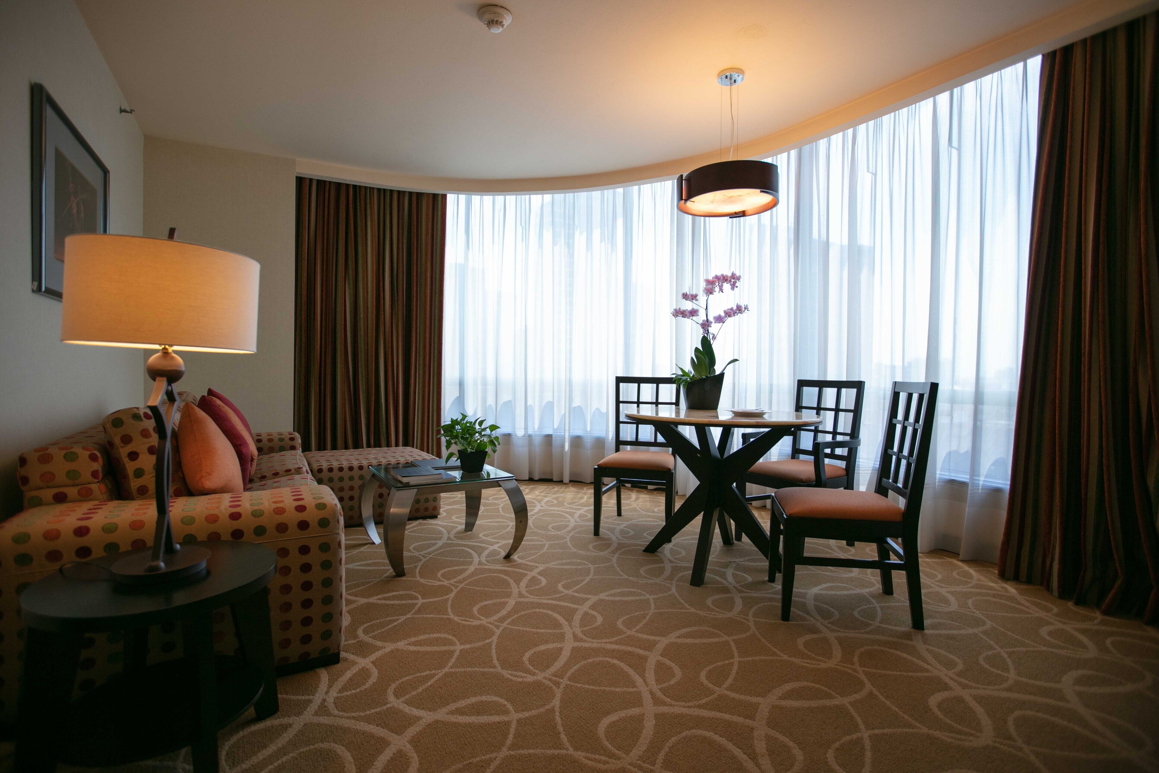 A hotel room with a sofa, a small table with a lamp, a dining table with chairs, and large windows with curtains.