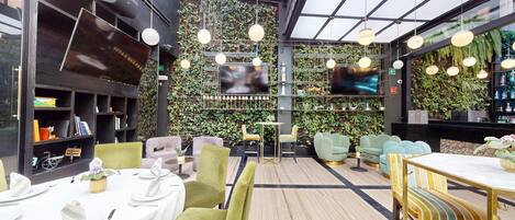 Un interior moderno con techo alto, pared verde con plantas y una zona de bar con televisor.