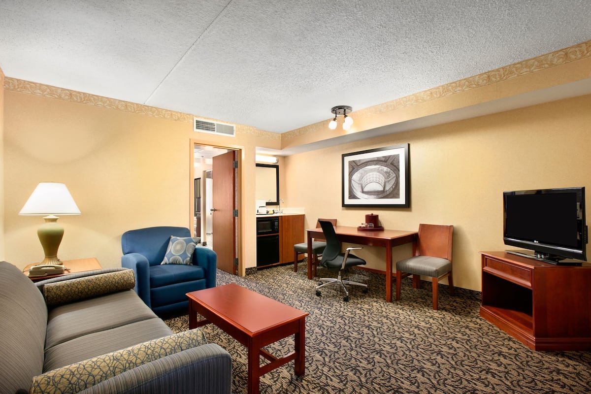 A hotel room with a sofa, a small table, a desk with a chair, a television, and a framed picture on the wall.