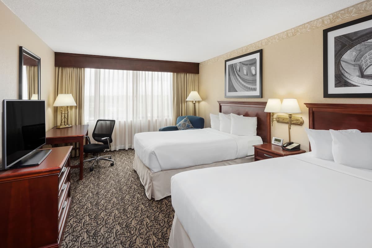 A hotel room with two beds, a desk with a TV, a chair, a desk lamp, a bedside table with a phone, and a large window with curtains.