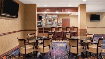 Un espace bar d’hôtel avec plusieurs tables rondes et des chaises, un comptoir de bar où sont exposées différentes bouteilles, et un téléviseur fixé au mur.