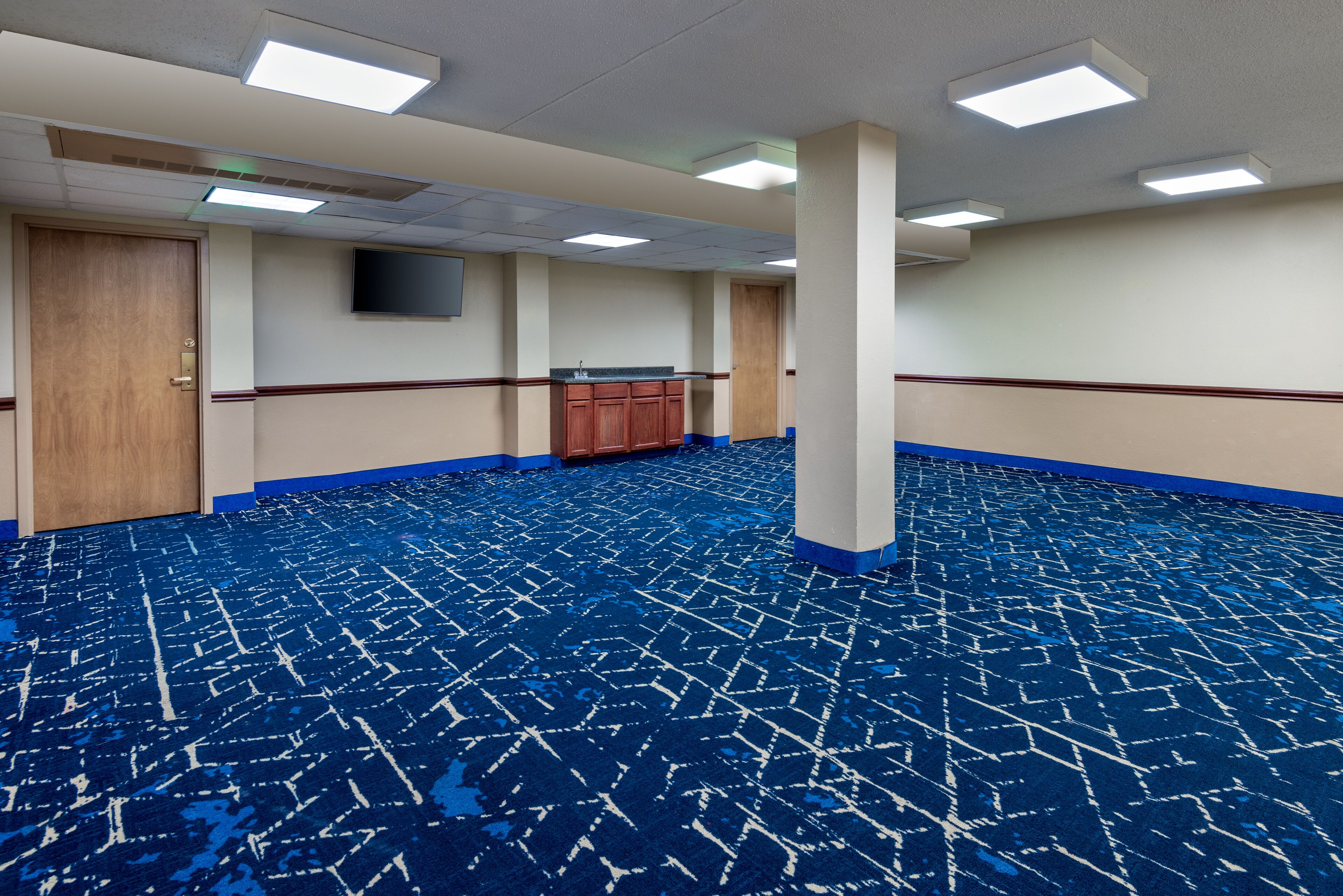 A large, empty room with a patterned blue carpet, wooden door, and a small kitchenette area.