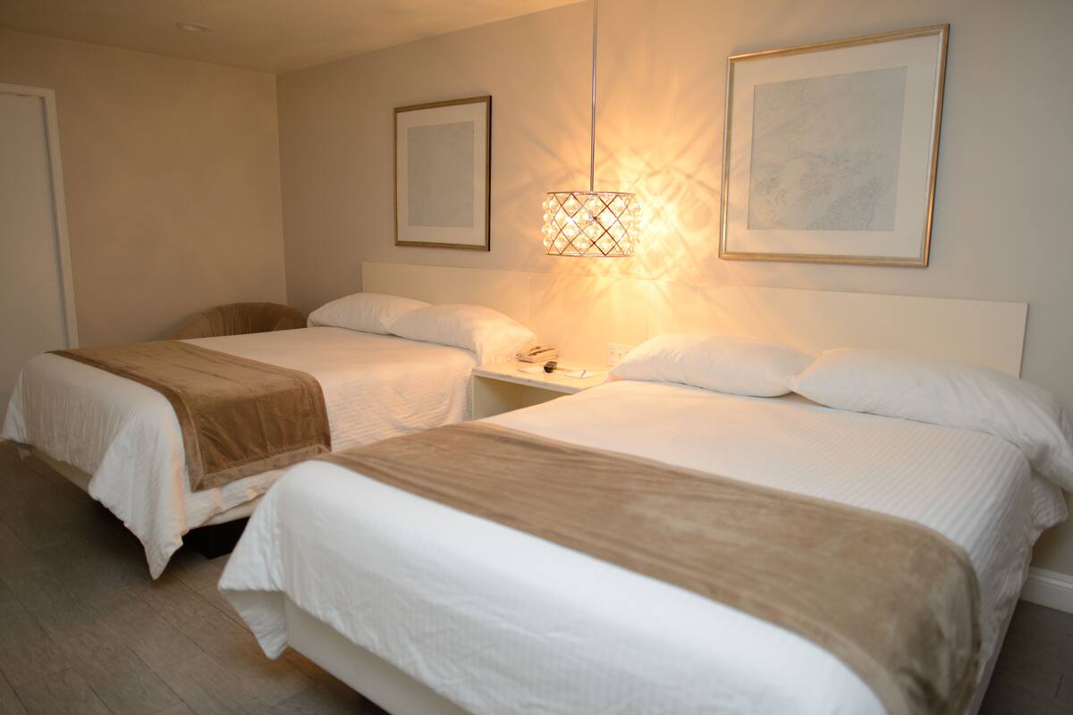 A hotel room with two beds, a bedside table, a wall-mounted lamp, and two framed pictures.
