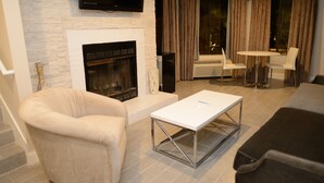 A living room with a fireplace, a flat-screen TV mounted above it, a white coffee table, and two white chairs around a small round table. - Hotel Le Rêve Pasadena (Pasadena)