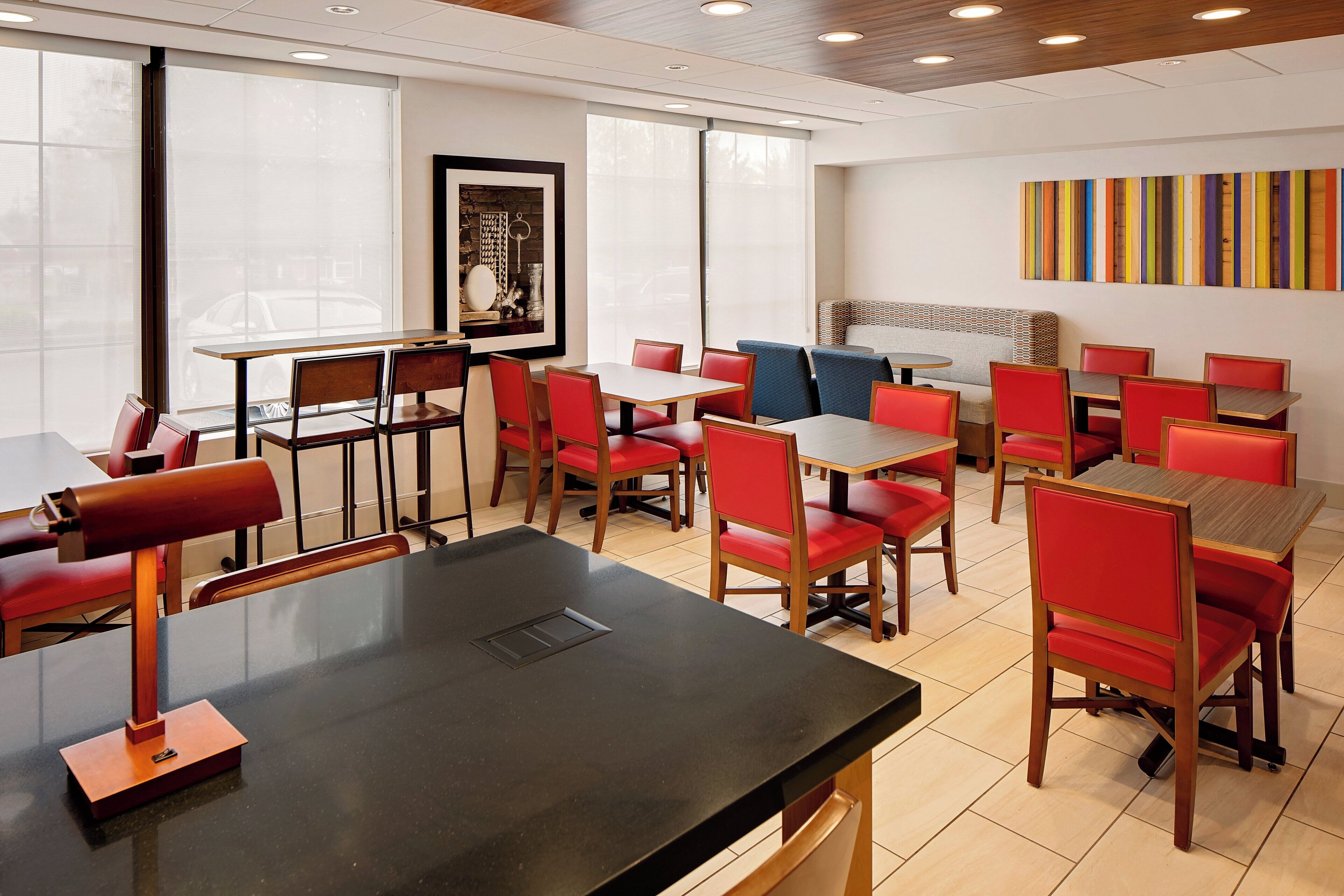 A modern dining area with red upholstered chairs, wooden tables, and a colorful abstract art piece on the wall.