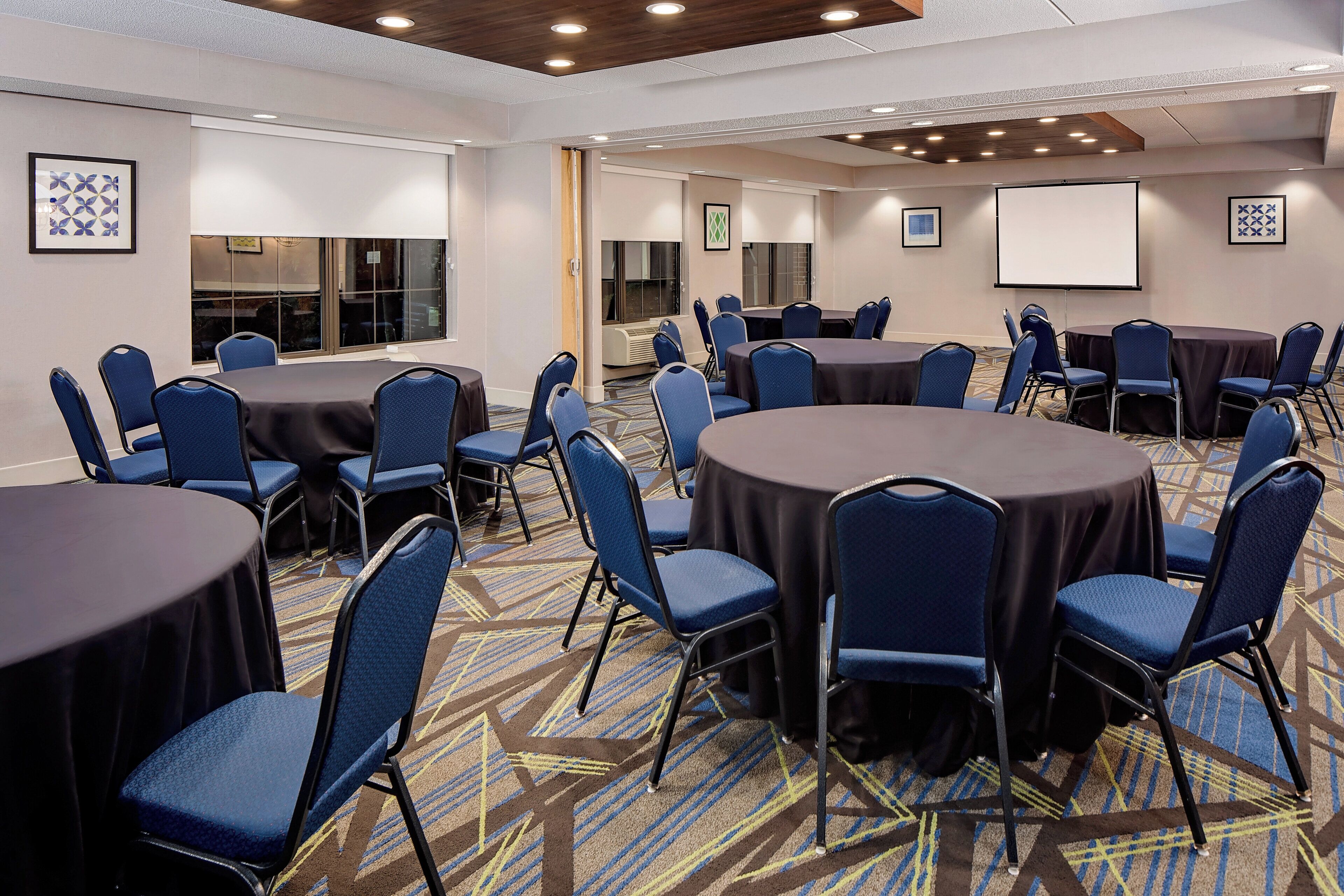Sala de conferências com mesas redondas e cadeiras azuis, piso coberto por carpete e tela de projeção.