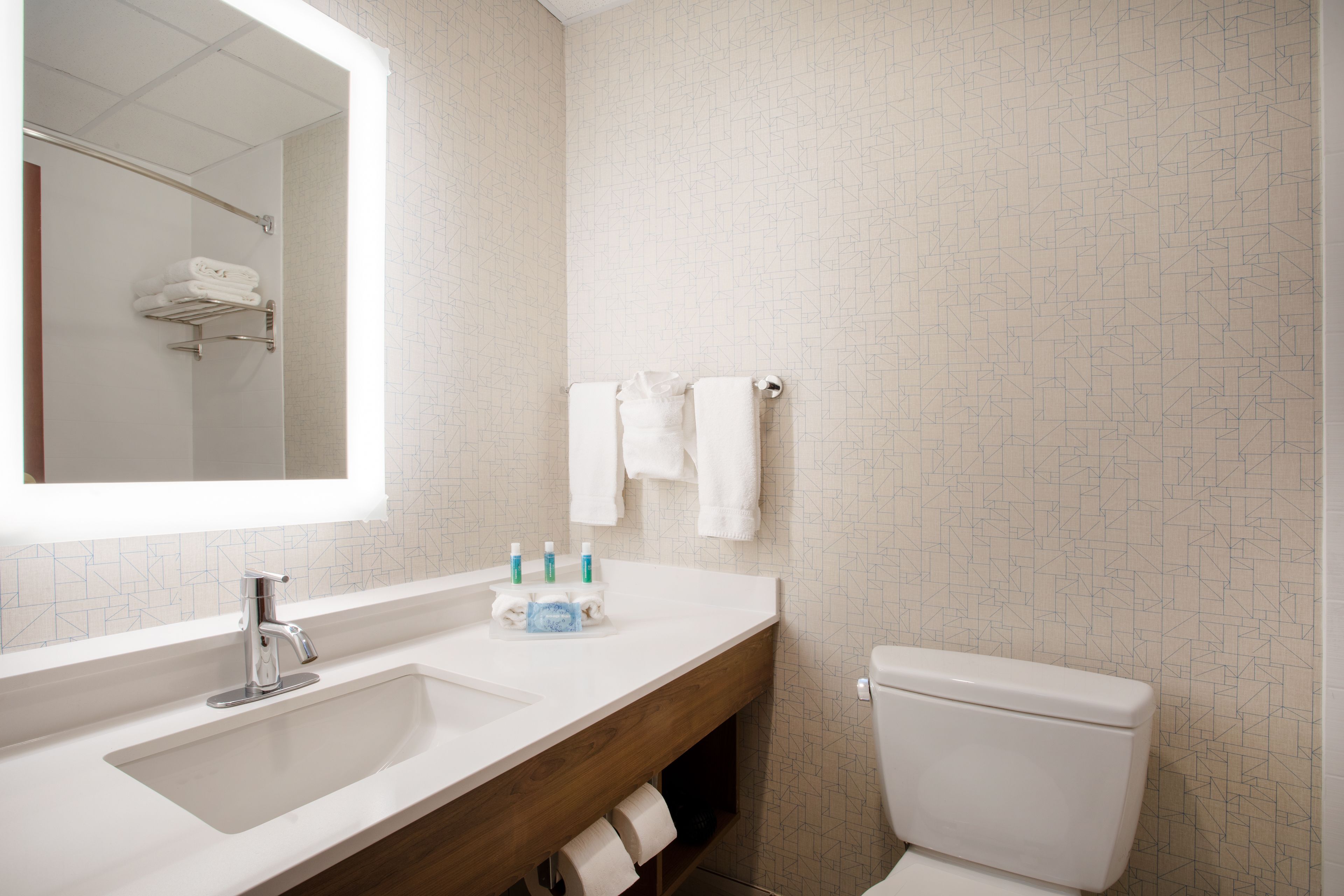 Un baño moderno con lavamanos, espejo y retrete blancos. Hay toallas y artículos de aseo en la encimera.