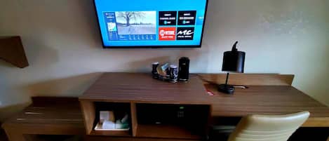A hotel room with a wooden desk, a flat-screen TV mounted on the wall, a desk lamp, and a chair.