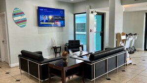 A modern reception area with a flat-screen TV displaying a welcome message, a black leather sofa, a small table with a vase, and a wall-mounted surfboard decoration.