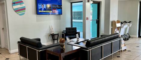 A modern reception area with a flat-screen TV displaying a welcome message, a black leather sofa, a small table with a vase, and a wall-mounted surfboard decoration.