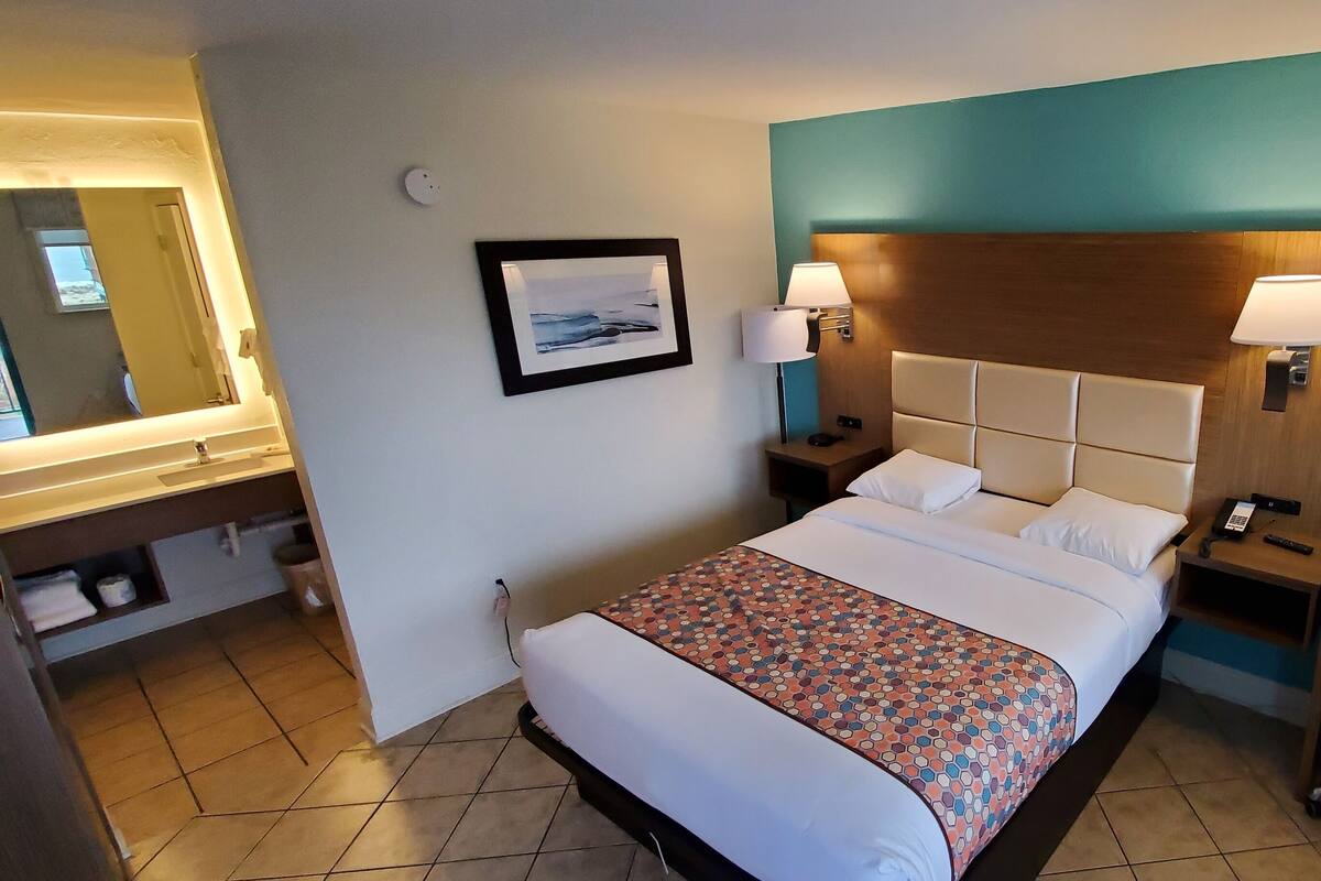 A hotel room with a bed, two bedside tables, a desk, a chair, a mirror, and a wall-mounted picture.