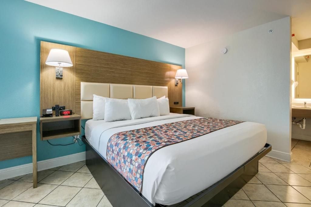 Quarto de hotel com cama, luminárias de cabeceira, mesa de cabeceira e relógio fixado na parede.
