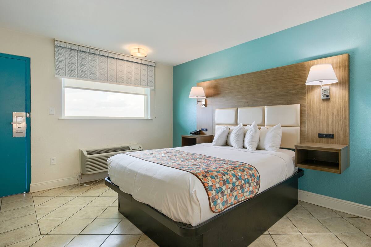 A modern hotel room with a large bed, a window with blinds, a wall-mounted lamp, and a built-in shelf.