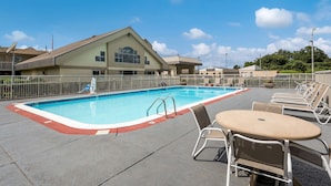 A swimming pool with lounge chairs and a table, surrounded by a fence, and a building with a sloped roof in the background.