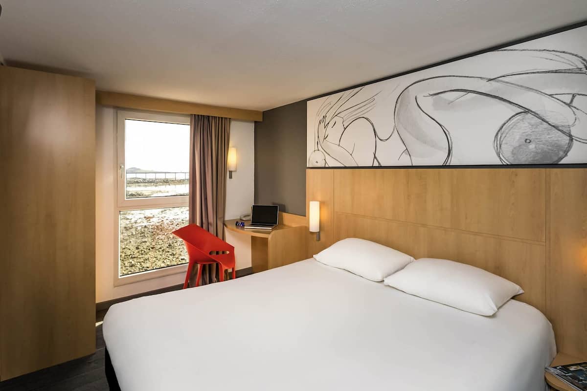 A hotel room with a large bed, a desk with a laptop, a red chair, and a view of the beach.