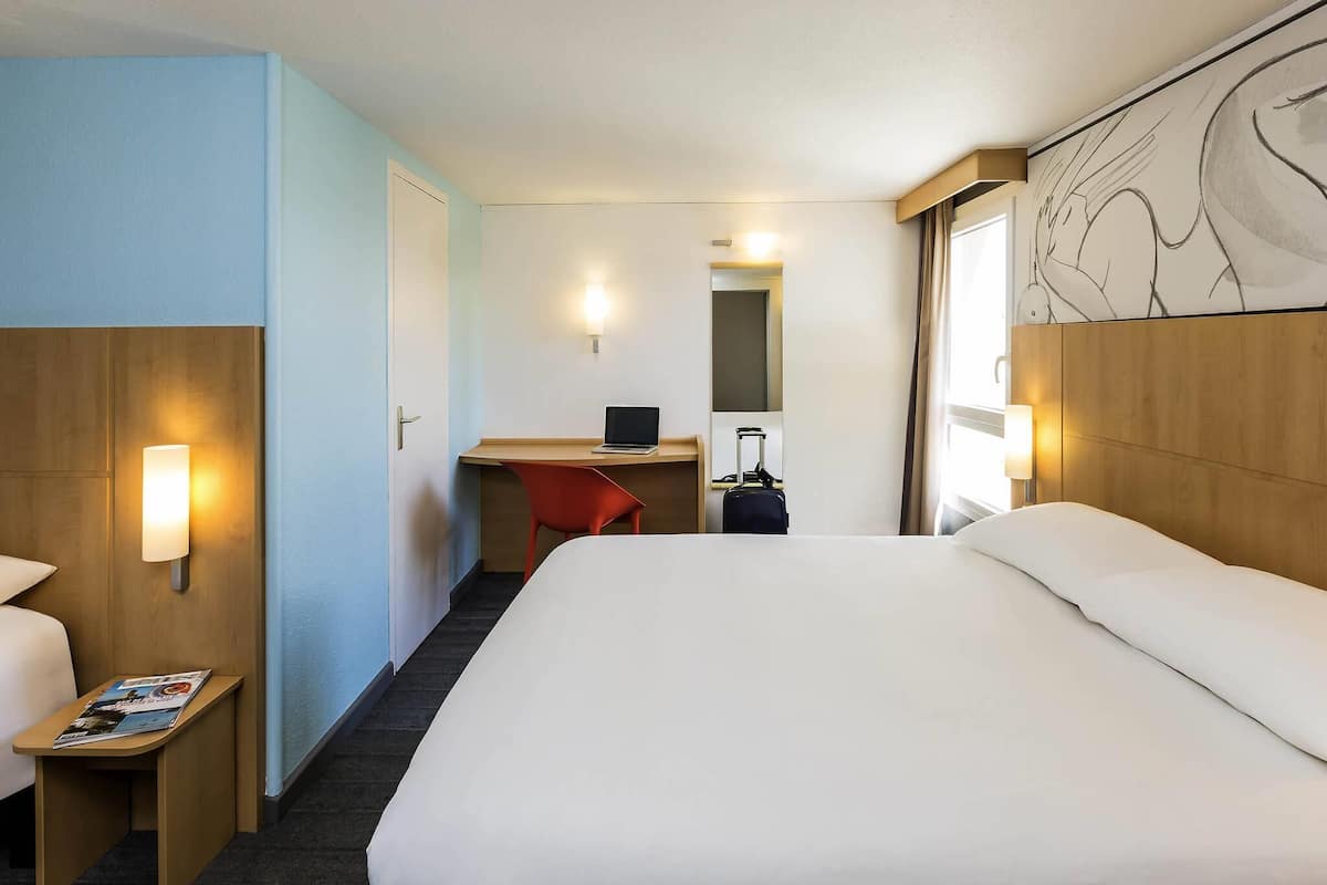 A hotel room with a bed, a desk, a chair, a small table with magazines, a suitcase, and a wall-mounted lamp.