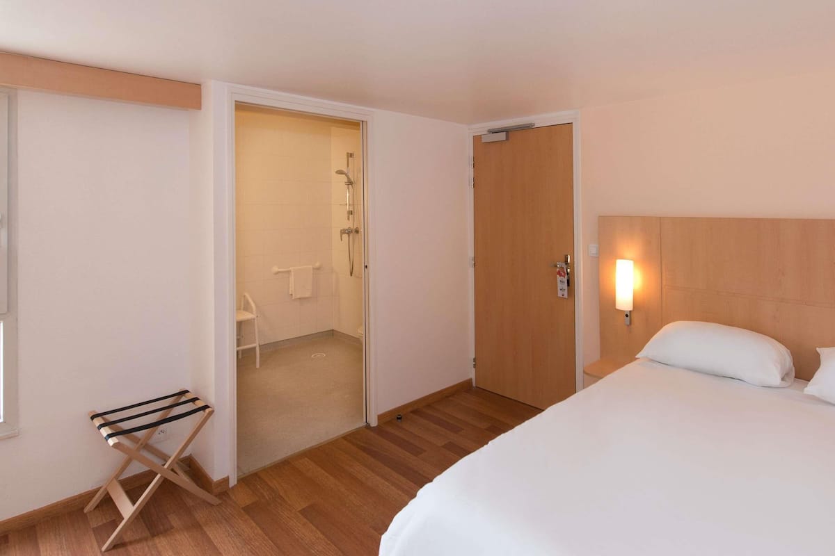 A hotel room with a bed, a wooden headboard, a nightstand, a wall-mounted light, a door, and a shower area.