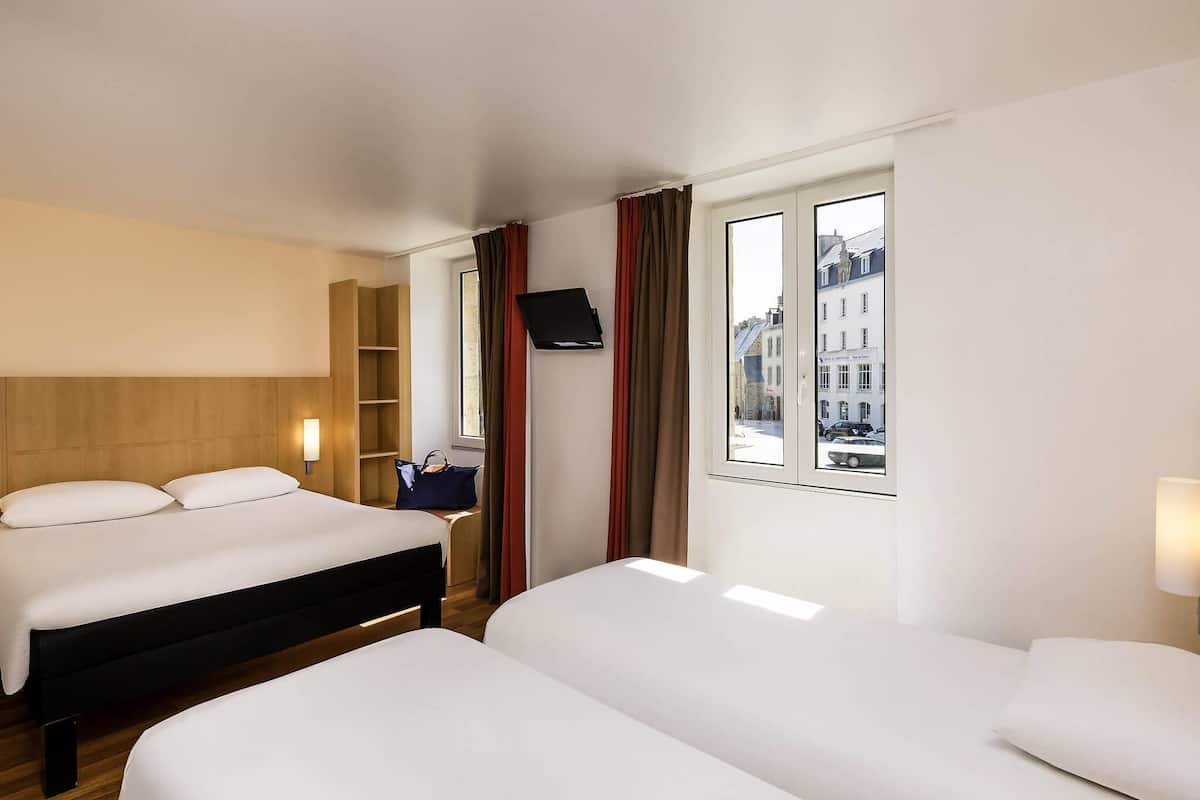 A hotel room with two beds, a wooden headboard, a shelf unit, a flat-screen TV, and a window with a city view.