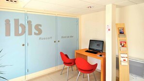 Una habitación con un separador de vidrio que indica 'ibis Roscoff' y 'Bord de Mer'. Hay un escritorio de madera con una computadora portátil, dos sillas rojas y un estante con folletos.