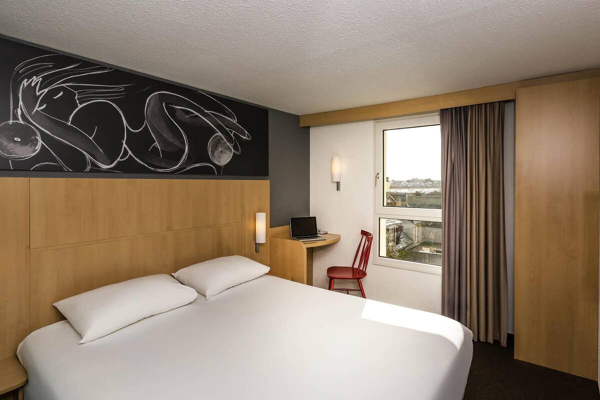 A hotel room with a large bed, a desk with a laptop, a red chair, a window with curtains, and a wall art piece.