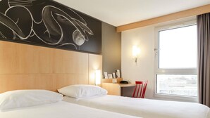 Habitación de hotel con dos camas, un cabecero de madera, un escritorio, una silla, un ventanal con cortinas y una pared con diseño de pizarra.