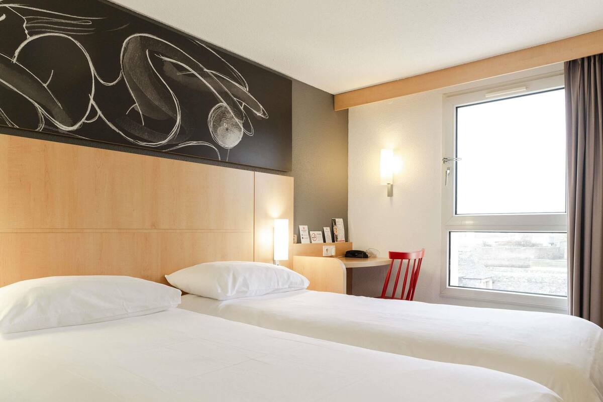 A hotel room with two beds, a wooden headboard, a desk, a chair, a window with curtains, and a wall with a chalkboard design.