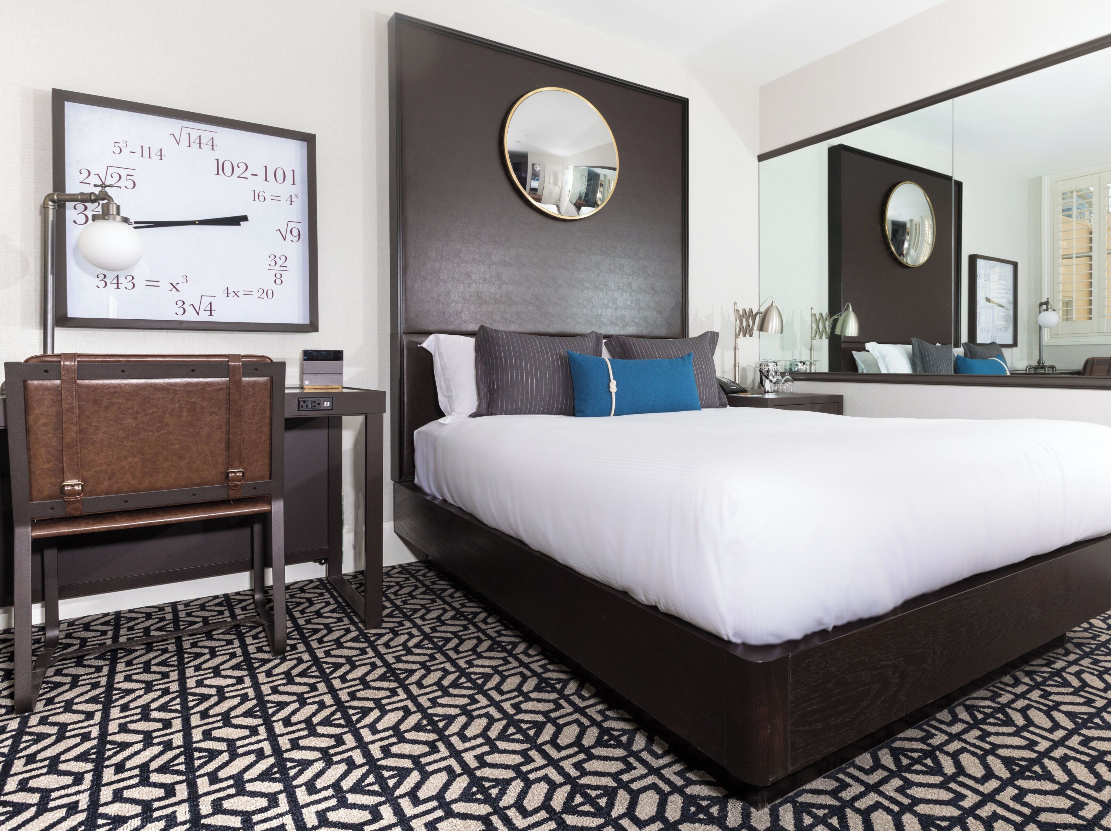 A modern hotel room with a large bed, a desk with a chair, a round mirror, and a wall clock displaying mathematical equations.