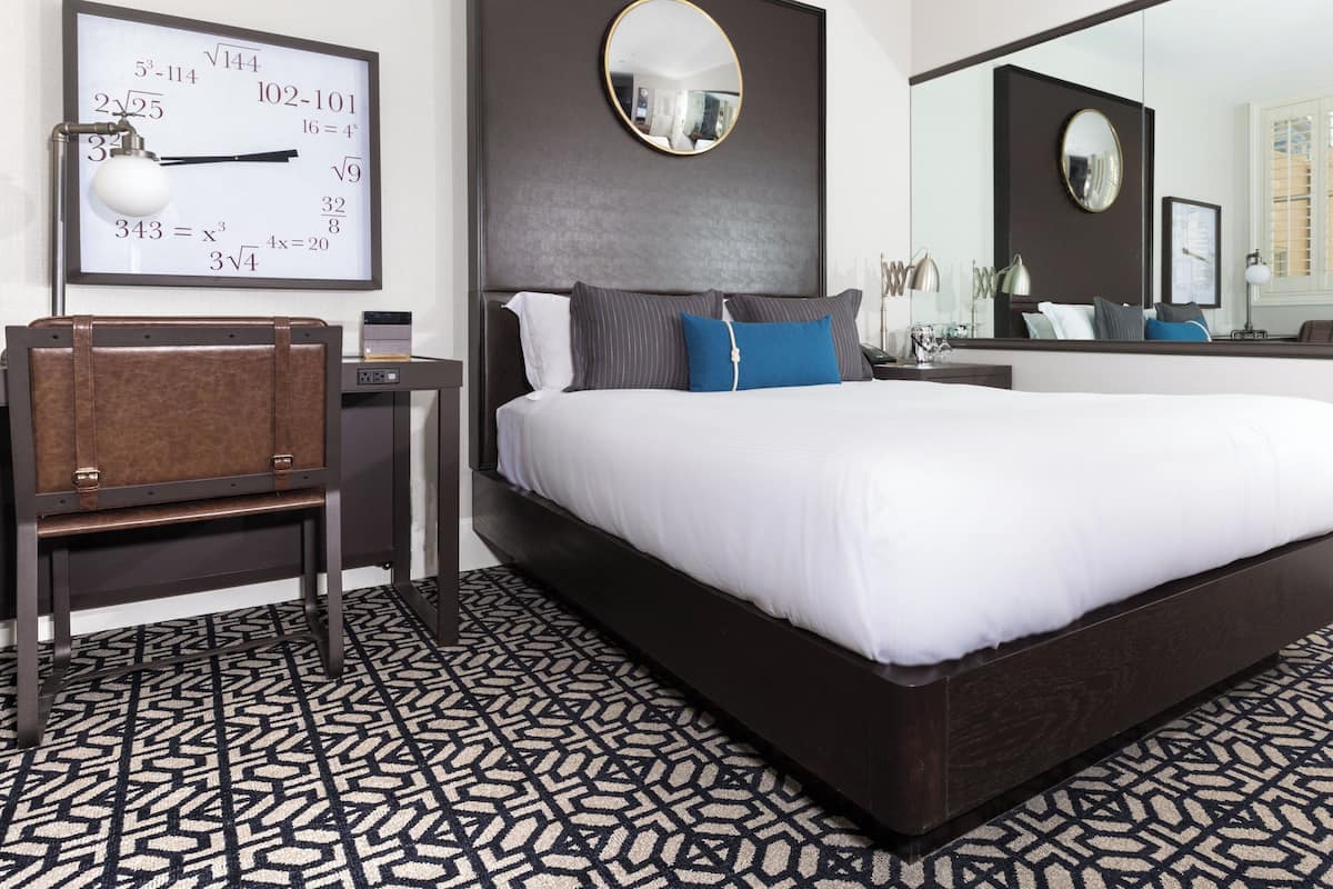 A modern hotel room with a large bed, a desk with a chair, a round mirror, and a wall clock displaying mathematical equations.
