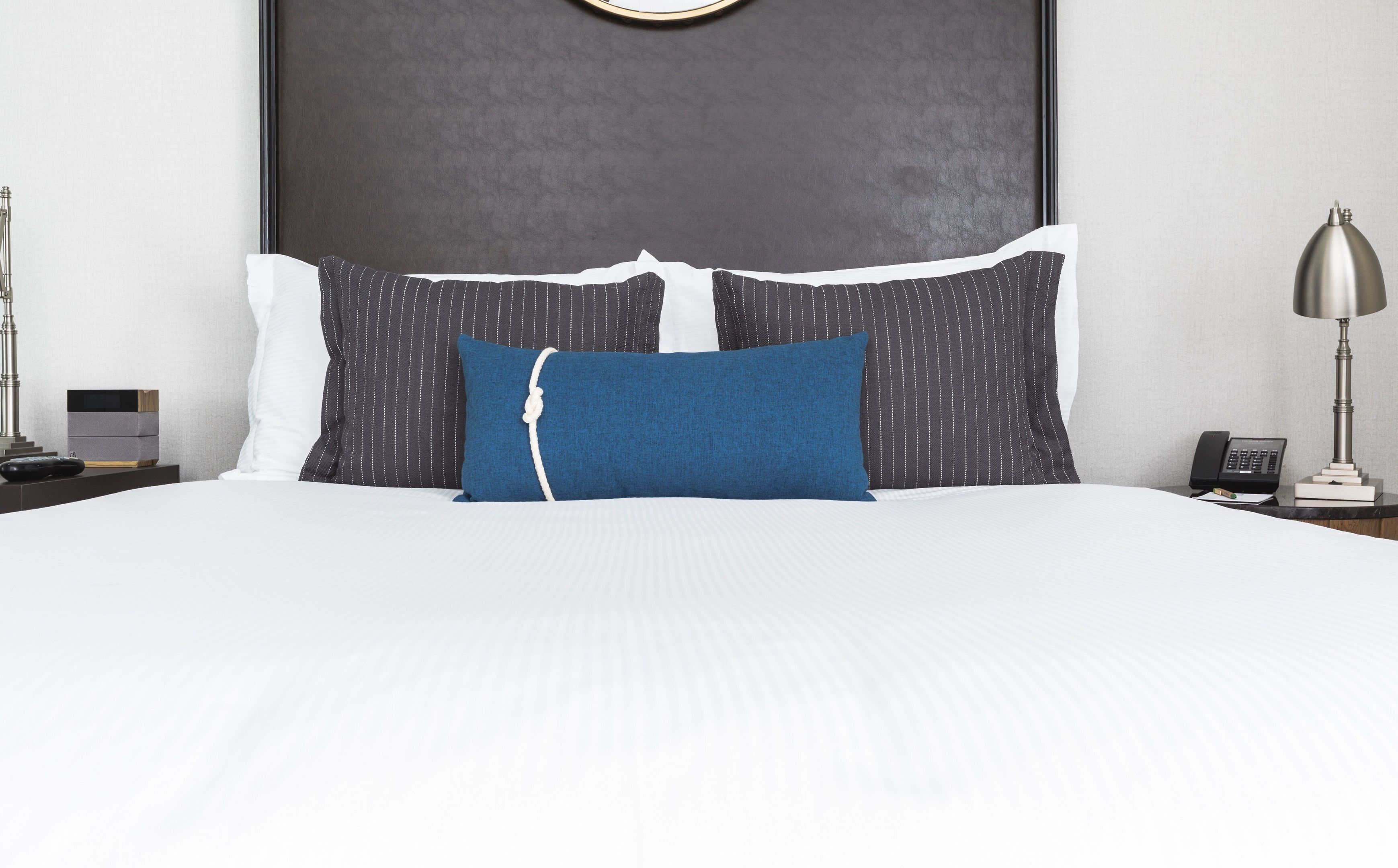 A neatly made bed with a white comforter and a blue accent pillow, a dark headboard, a bedside table with a lamp, and a small bedside table with a phone and a book.