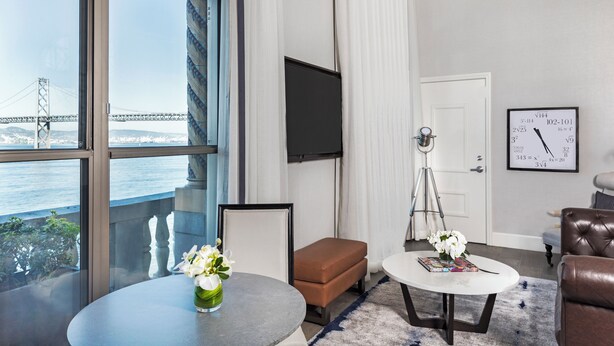 A modern living room with a large window overlooking a bridge, a flat-screen TV, a clock, and a brown leather sofa. - Harbor Court Hotel (San Francisco)