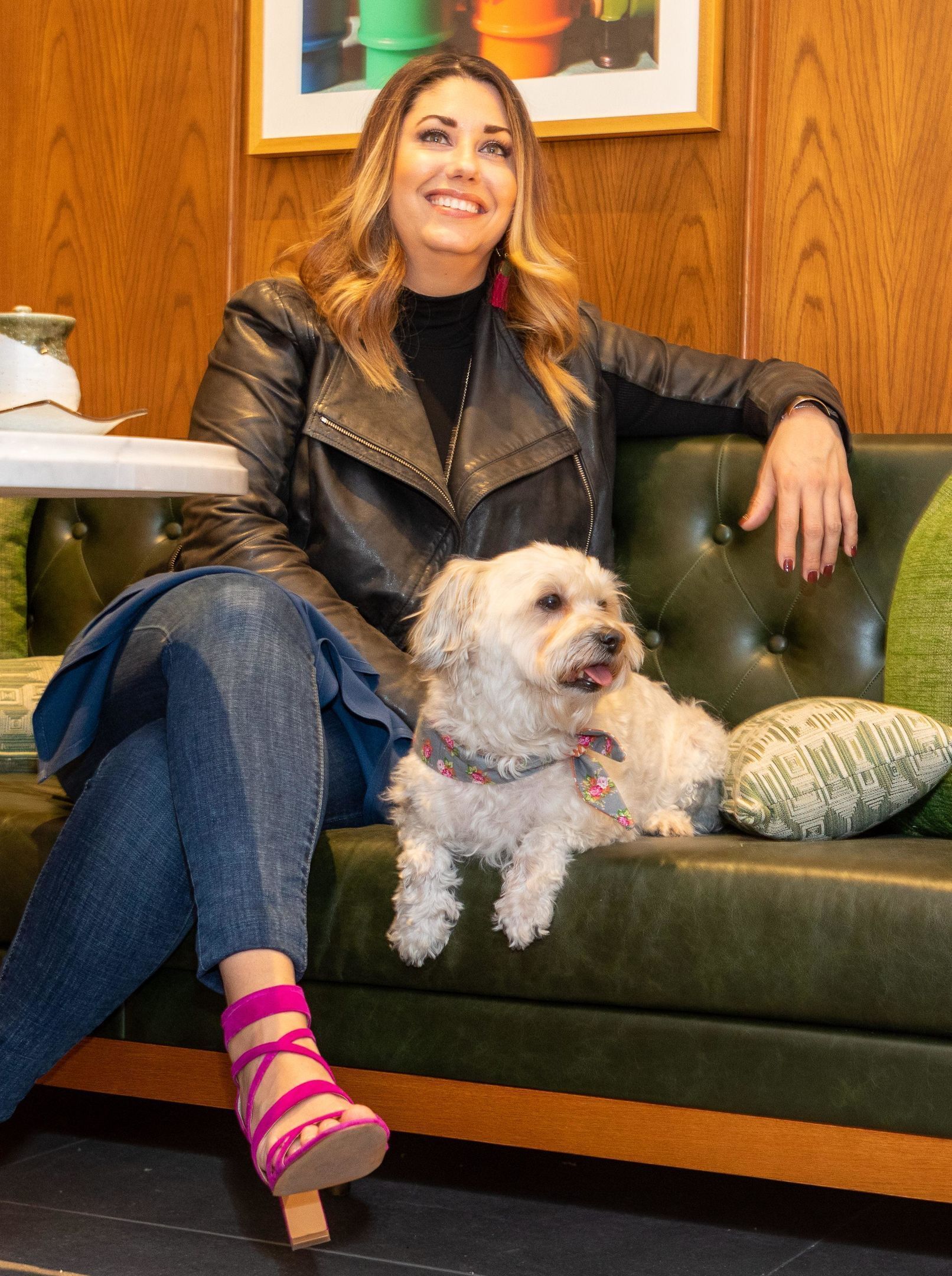 A person sitting on a green leather couch with a small white dog, wearing a leather jacket and jeans, with a wooden wall and a framed picture in the background.