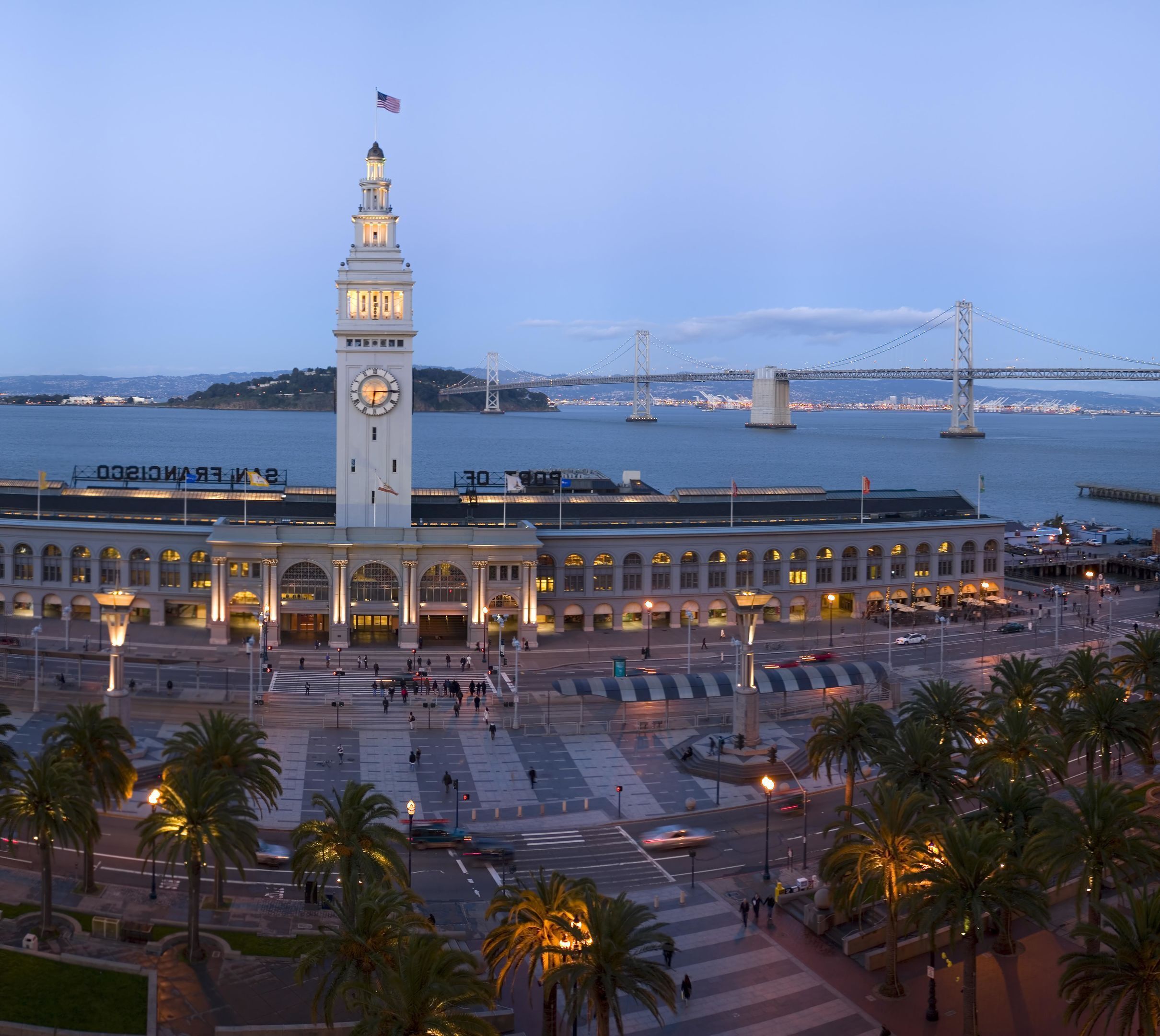 A large public square with a prominent clock tower, a bridge in the background, and palm trees.
