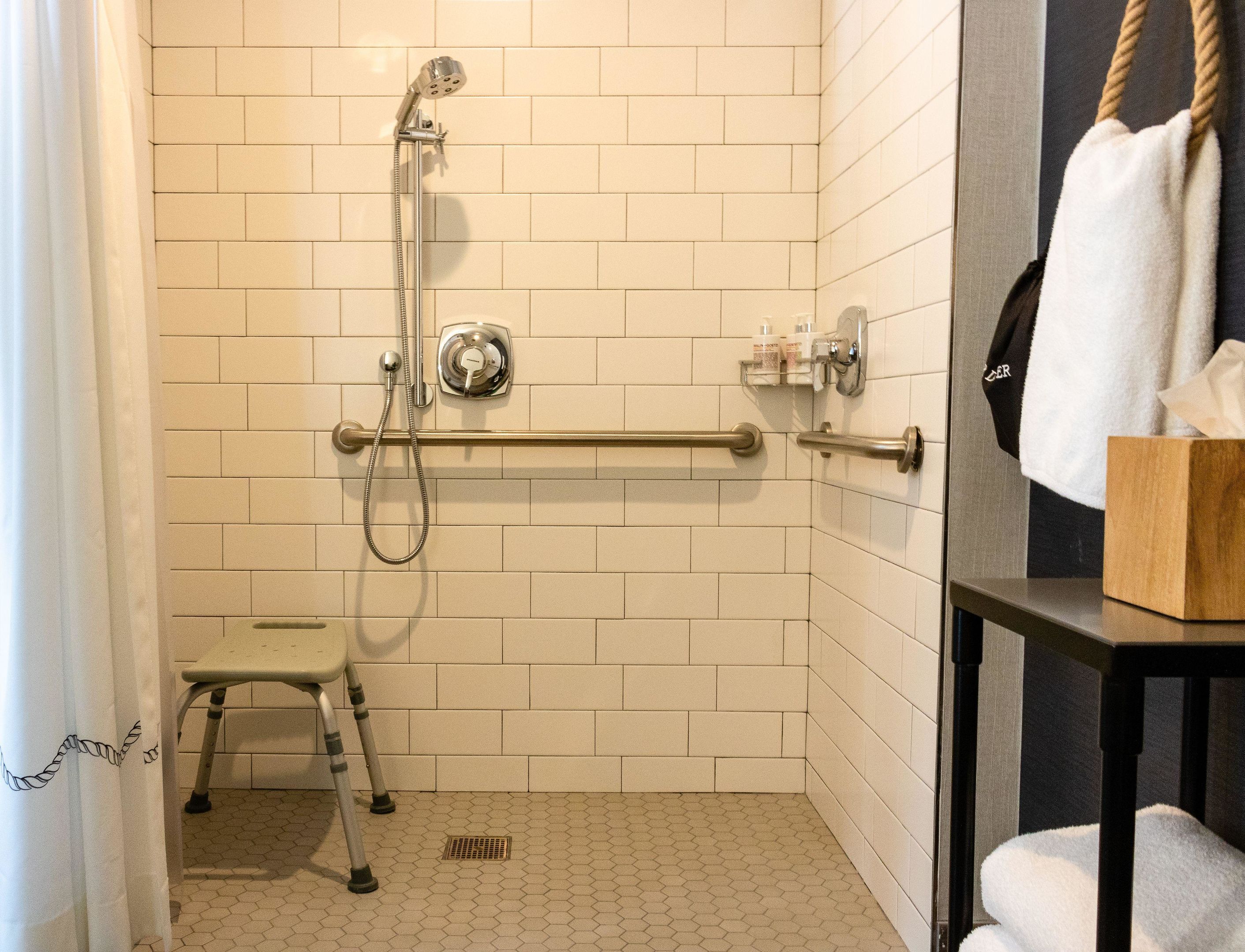Une salle de bain avec des murs carrelés de blanc, une douche équipée d’un pommeau de douche à main, d’une barre d’appui, d’un petit marchepied et d’un porte-serviettes avec une serviette.