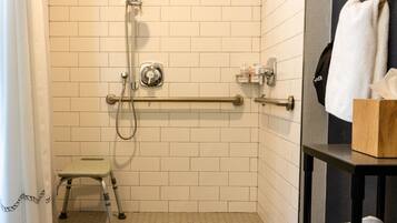 Une salle de bain avec des murs carrelés de blanc, une douche équipée d’un pommeau de douche à main, d’une barre d’appui, d’un petit marchepied et d’un porte-serviettes avec une serviette.