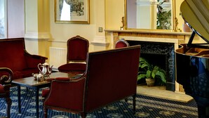 Una sala elegante con un piano, sillones tapizados en rojo, una mesa con tetera y una chimenea.