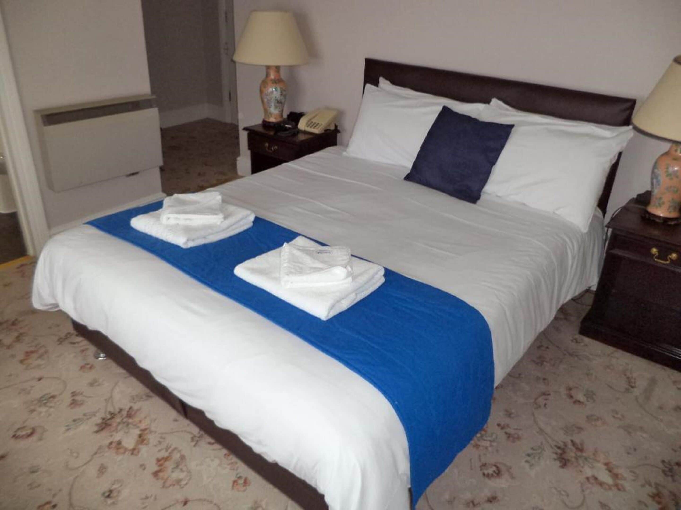 Una cama bien hecha con ropa de cama blanca y azul, dos toallas dobladas, una lámpara de noche y un teléfono en una mesita de noche.
