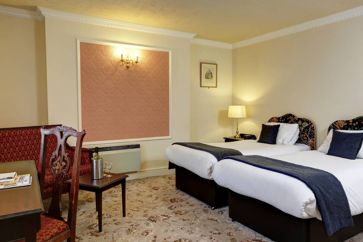 A hotel room with two beds, a wooden chair, a table with a lamp, and a framed picture on the wall.