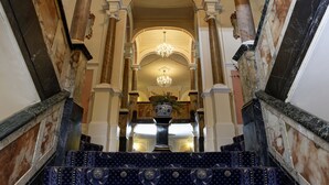 Una imponente escalera con columnas de mármol y un candelabro, que conduce a un espacio con techos altos y un gran jarrón sobre un pedestal.