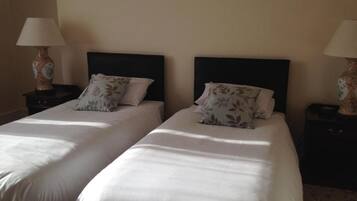 Quarto de hotel com duas camas de solteiro, cada uma com cabeceira escura e almofadas com padrão floral. Possui duas mesas de cabeceira com luminárias e um vaso decorativo em cada uma.